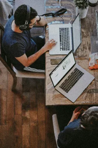 Person mit Kopfhörern arbeitet an zwei Laptops an einem Holztisch, von oben fotografiert