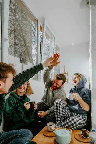 Vier lachende Menschen sitzen mit Kaffeetassen am Fenster und geben sich ein High-Five